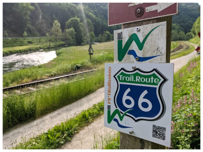 Wanderzeichen für die Trail.Route 66 an einem Holzpfahl neben einem Weg, Bahngleisen und Fluss
