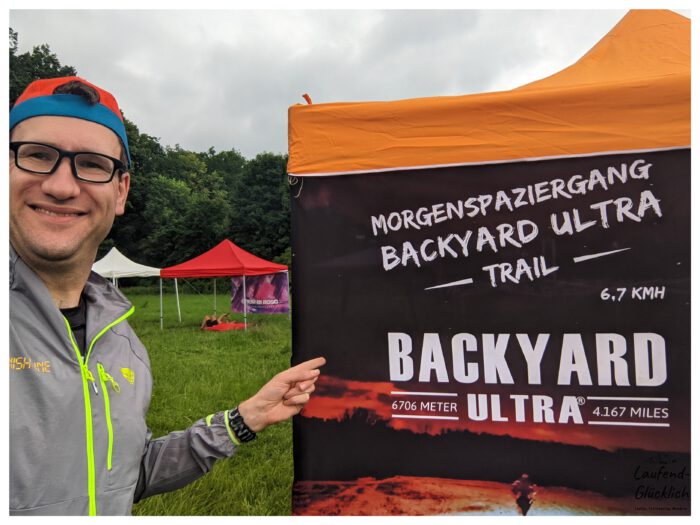 Ein lächelnder Läufer Thorsten zeigt auf ein Banner des „Morgenspaziergang Backyard Ultra Trail“