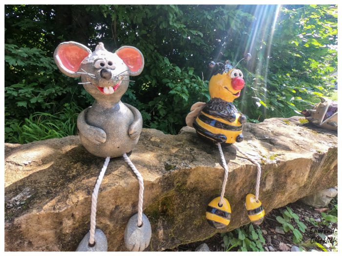 Zwei Keramikfiguren einer Maus und einer Biene mit Schlenkerbeinen sitzen auf einer Steinmauer im Sonnenlicht