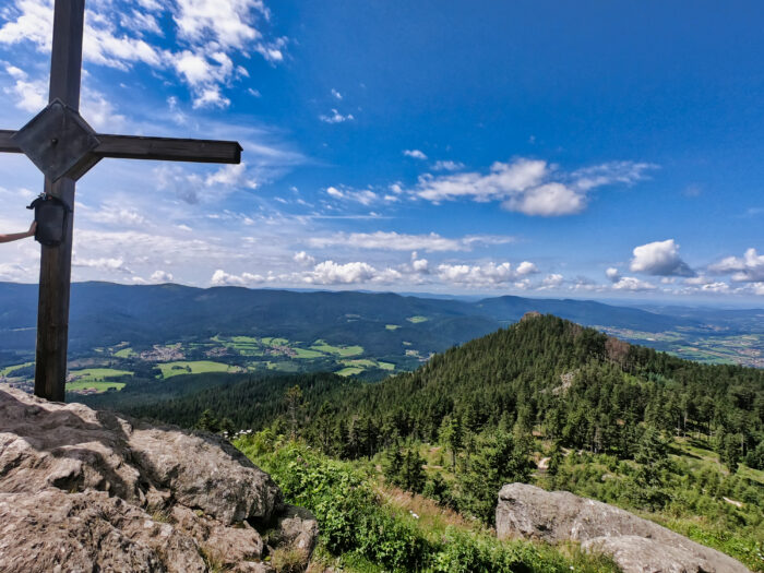 Gipfelkreuz auf einem Felsen mit Panoramablick über eine bewaldete Berglandschaft unter blauem Himmel