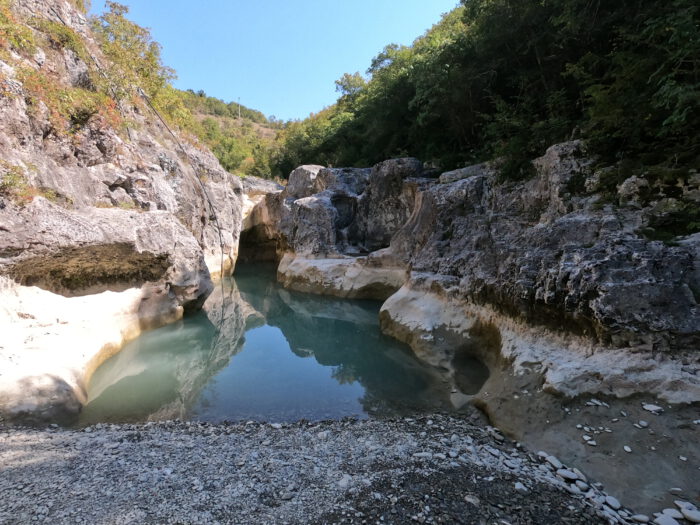 Ein türkisgrünes Wasserbecken in einer felsigen Schlucht, umgeben von hellen Kalksteinen und bewaldeten Hängen