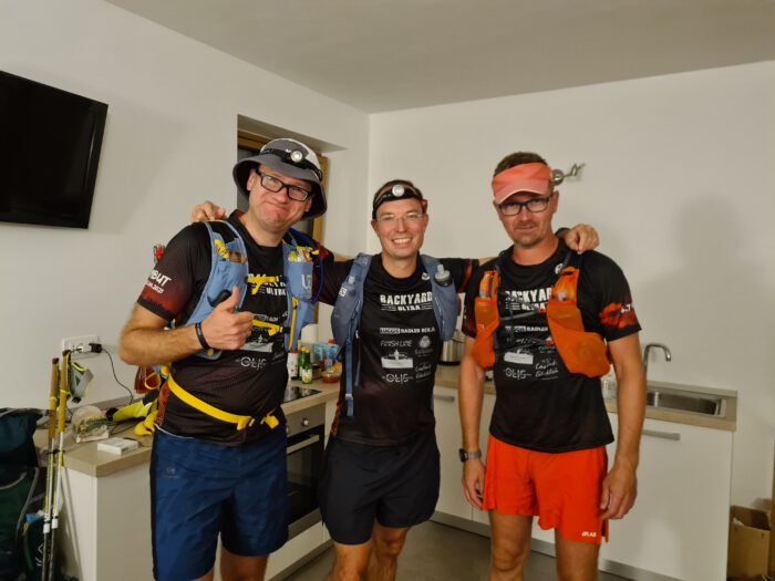 Drei Läufer in Trailrunning-Ausrüstung mit Stirnlampen und Trinkwesten posieren gemeinsam in einer Küche