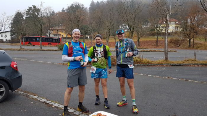 Drei Läufer in Sportkleidung, unter anderem Ingo und Thorsten, mit Startnummern stehen lächelnd mit Bierflaschen auf einem Parkplatz
