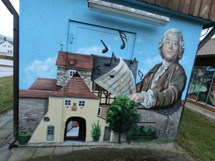 Wandgemälde eines Komponisten mit Perücke und Notenblatt neben historischen Fachwerkhäusern und einem Torbogen