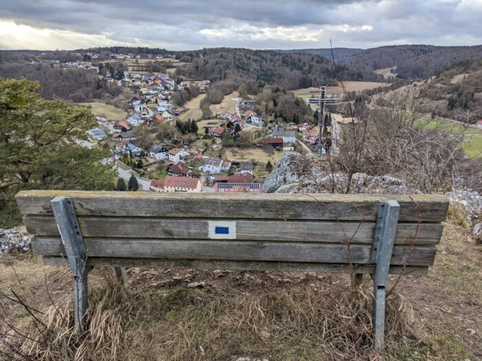 Blick von einer Holzbank auf ein Dorf im Tal, umgeben von bewaldeten Hügeln, mit einem Gipfelkreuz auf einem Felsen