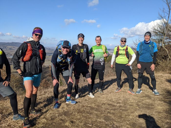Sechs Trailrunner posieren lächelnd auf einer Anhöhe vor einer weiten Tallandschaft bei sonnigem Wetter