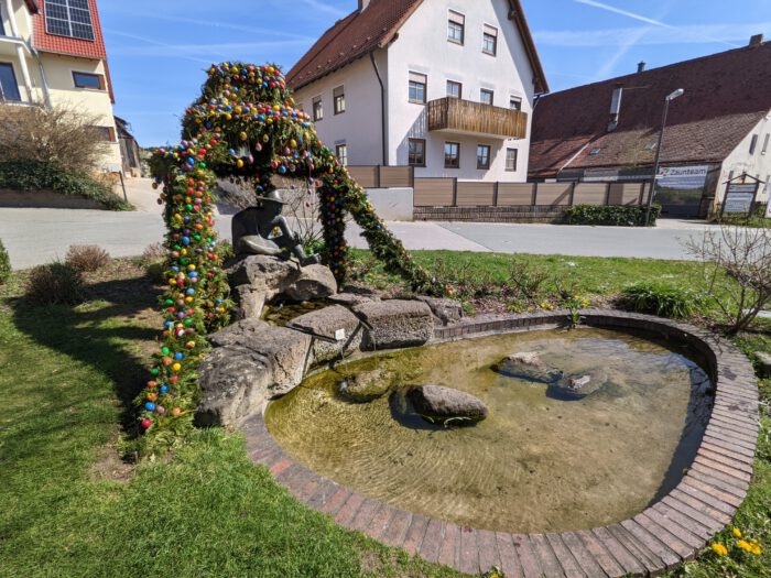 Mit bunten Ostereiern und Tannenzweigen geschmückter Osterbrunnen mit einer Bronzestatue an einem kleinen Teich