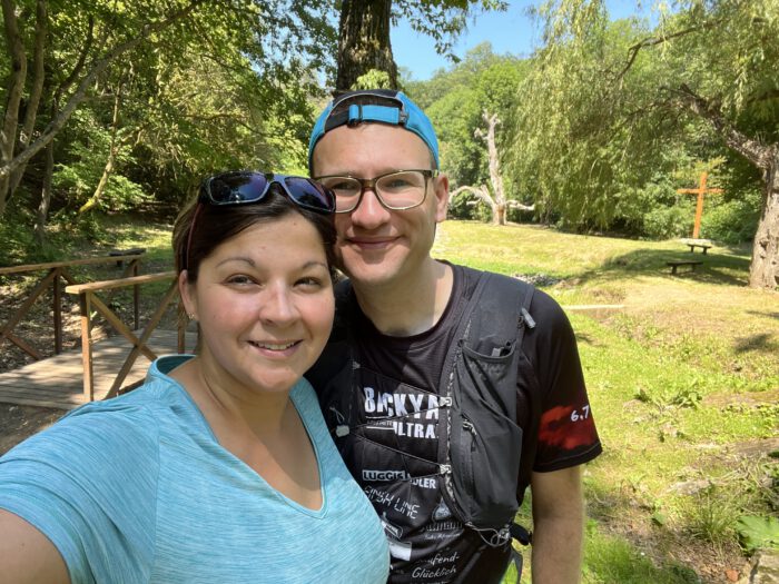 Ein lächelndes Paar (Mareike und Thorsten) macht ein Selfie in einer sonnigen Waldlandschaft mit einer kleinen Holzbrücke im Hintergrund