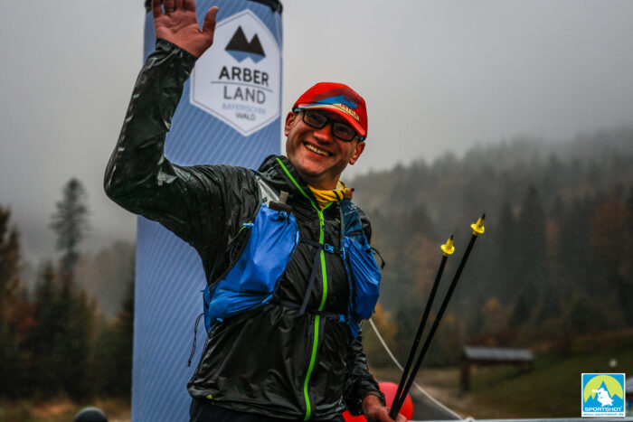 Lächelnder Trailrunner winkt vor einem Arber Land Banner im Bayerischen Wald