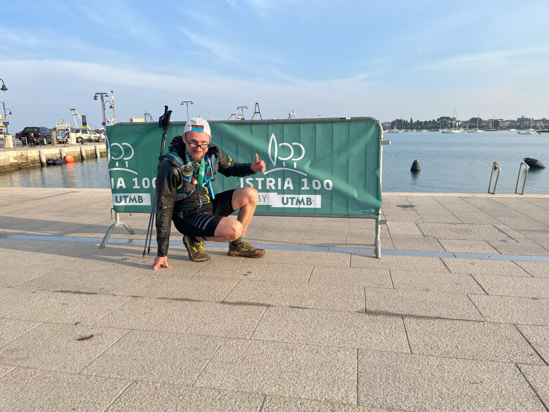 Thorsten hockt mit gehobenem Daumen vor einem grünen Banner mit der Aufschrift Istria 100 by UTMB an einer Hafenpromenade in Umag nach dem Zieleinlauf
