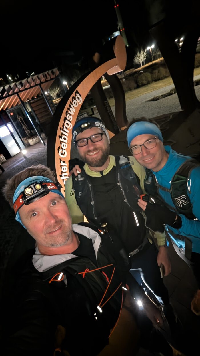 Drei Läufer, Flo, Thorsten und Hansi, mit Stirnlampen und Laufwesten machen nachts ein Selfie vor einem Schild zum Gebirgsweg