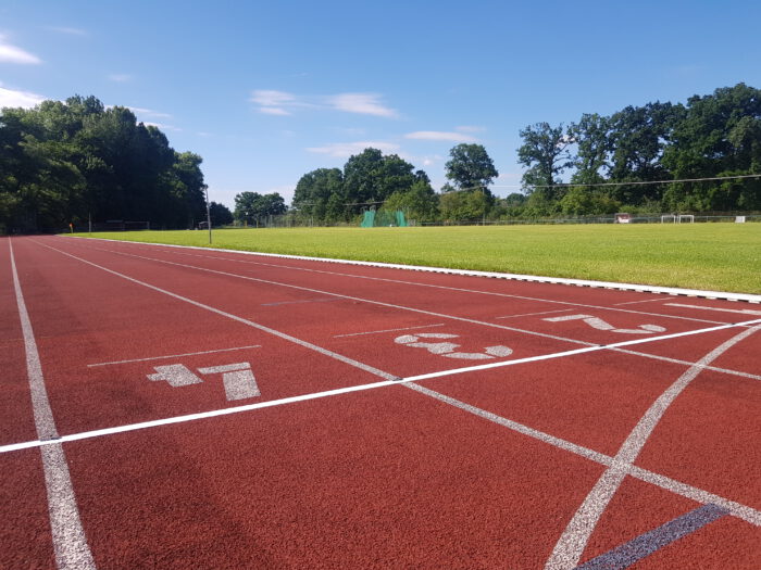 Rote Laufbahn auf einem Sportplatz mit weißen Markierungen und Bäumen im Hintergrund