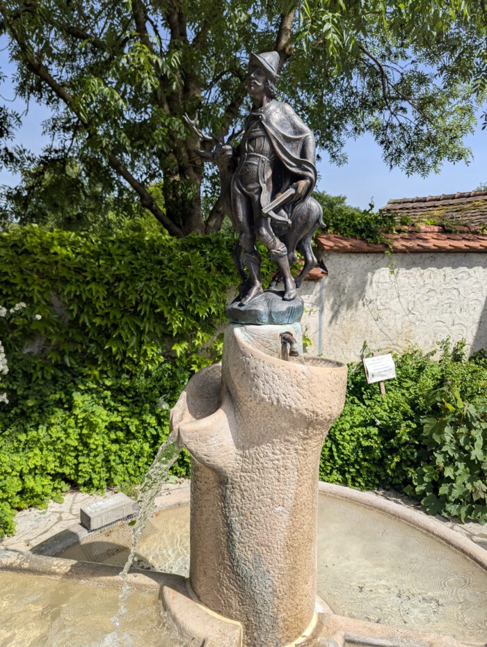 Steinbrunnen mit einer Bronzestatue eines Jägers mit Hirsch und Armbrust, aus dem Wasser in ein Becken fließt