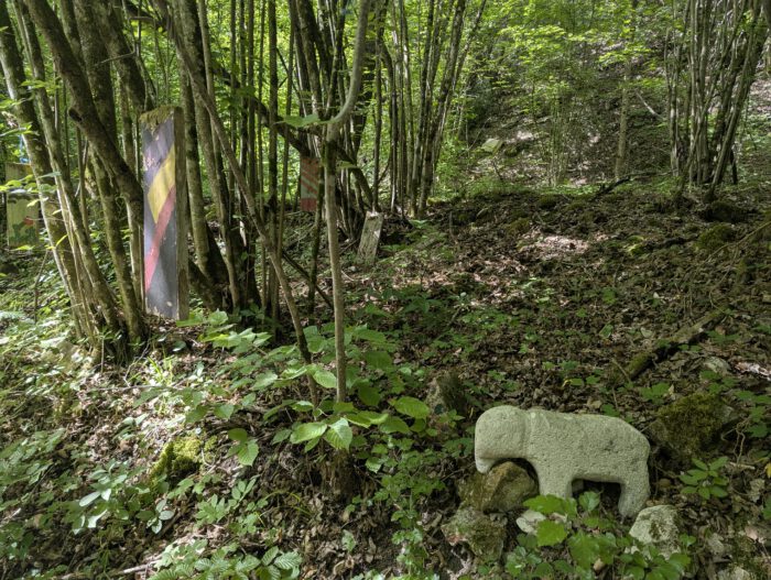 Steinfigur eines Elefanten am Boden und bemalte Holztafeln an Bäumen in einem schattigen Waldstück
