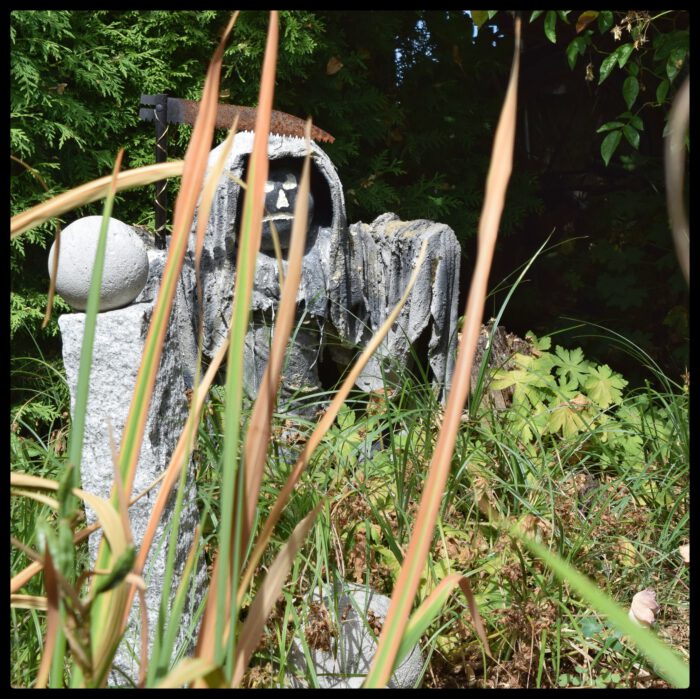 Eine mystisch wirkende Figur in einem grauen Kapuzengewand mit einer rostigen Sense inmitten von Pflanzen