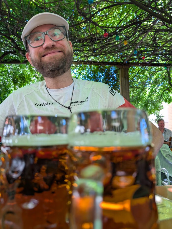 Ein lächelnder Mann mit Brille und Kappe sitzt hinter zwei gefüllten Bierkrügen im grünen Biergarten
