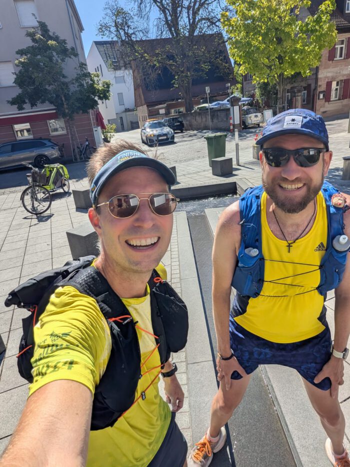 Zwei lächelnde Männer, Hansi und Thorsten, in Laufbekleidung mit Trinkwesten und Sonnenbrillen auf einem gepflasterten Platz