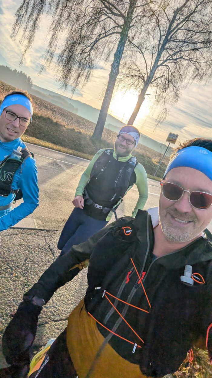 Drei Männer in Sportkleidung und mit Stirnbändern lächeln bei Sonnenaufgang auf einer Landstraße