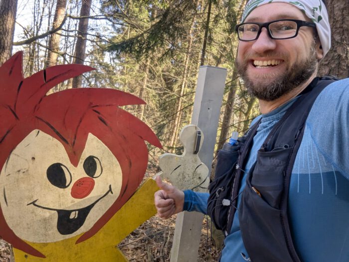 Ein lächelnder Mann in Wanderkleidung gibt einen Daumen hoch neben einer hölzernen Pumuckl-Figur im Wald