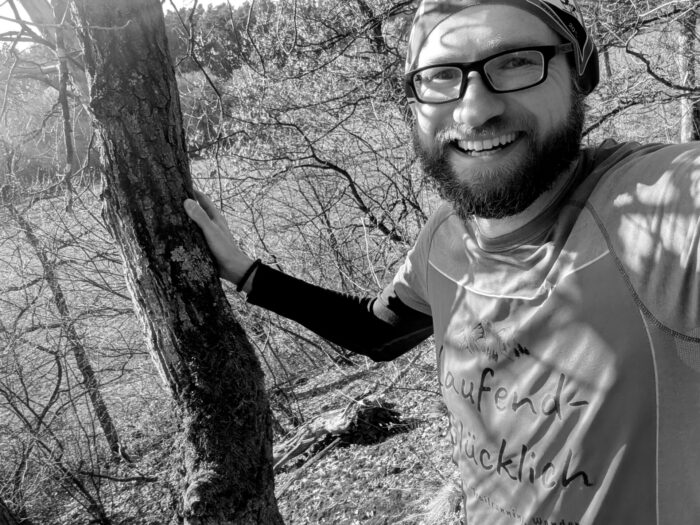 Ein lächelnder Mann mit Bart und Brille im Laufend-Glücklich-Shirt lehnt draußen an einem Baum