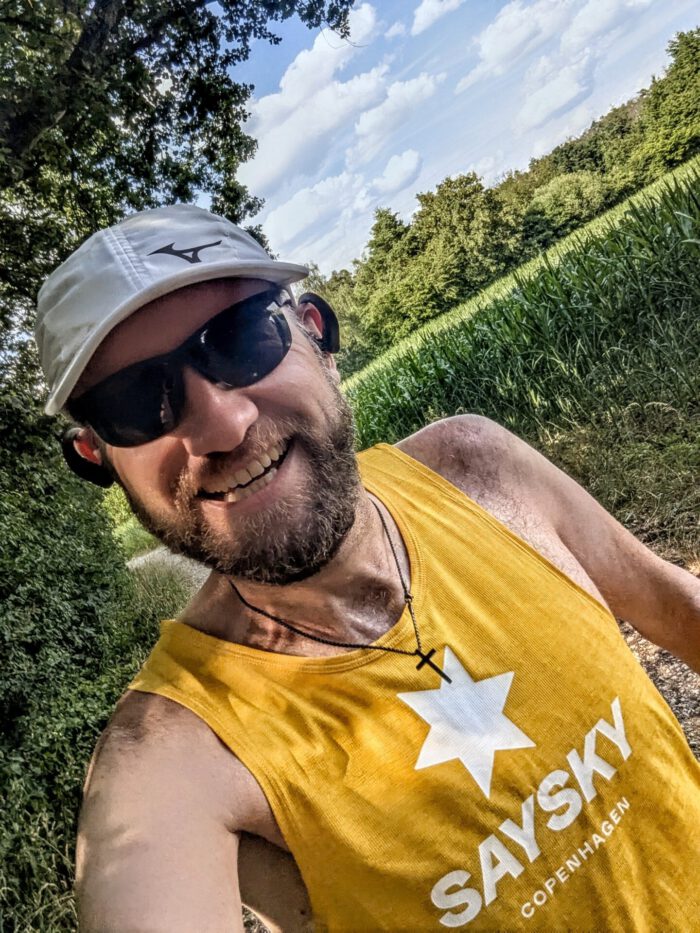 Lächelnder, verschwitzter Thorsten mit Kappe und Sonnenbrille in gelbem Sportshirt vor sommerlicher Landschaft