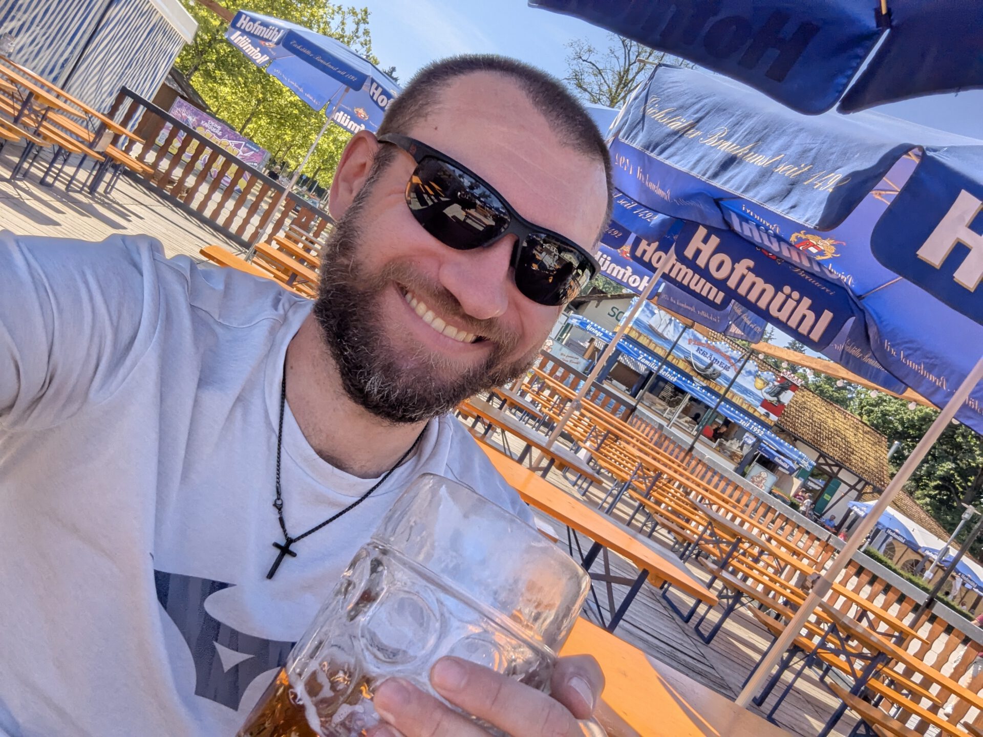 Ein lächelnder Mann mit Bart und Sonnenbrille hält in einem Biergarten eine Maß Bier