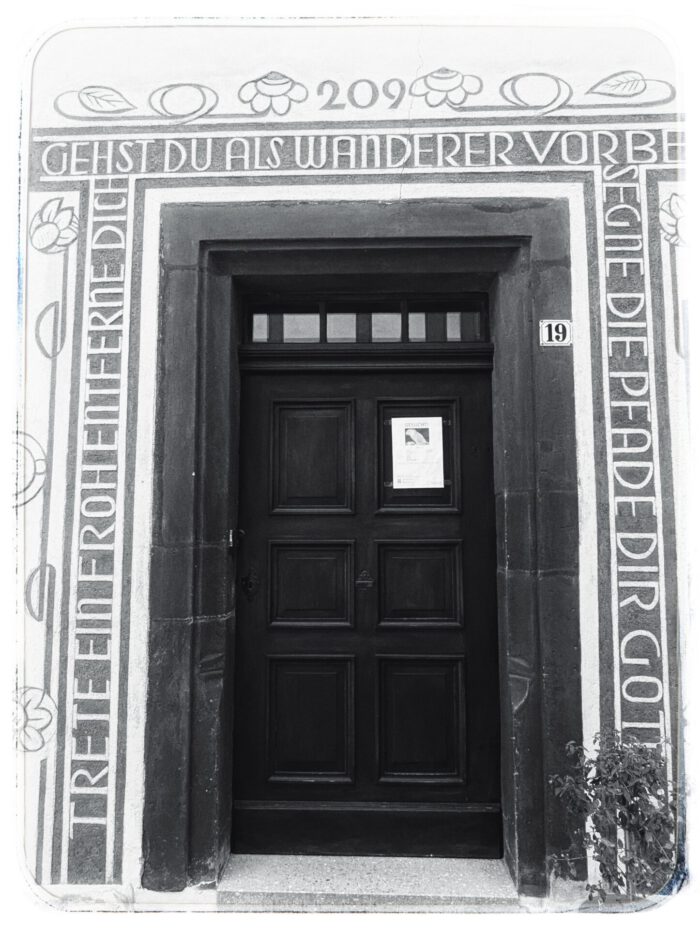Verzierte Tür mit der Aufschrift: Gehst du als Wanderer vorbei, trete ein, froh entferne dich, segne die Pfade dir Gott