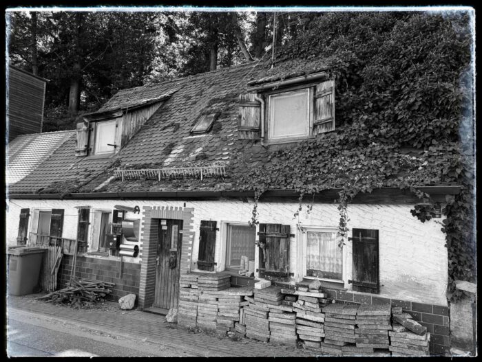 Altes kleines Haus mit Holzklappläden, teils efeuüberwachsenem Ziegeldach und gestapelten Gehwegplatten davor