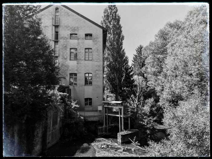 Historisches Mühlengebäude an einem Flusslauf mit kleiner Wehranlage und umgebenden Bäumen