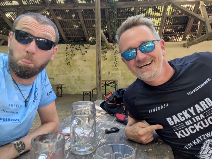 Zwei Läufer in Sportbekleidung und Sonnenbrillen sitzen gut gelaunt an einem Holztisch im Biergarten
