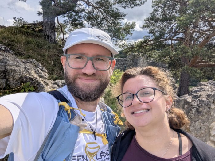 Ein lächelndes Paar, Thorsten mit Mel, mit Brillen macht ein Selfie im Freien vor felsiger Naturkulisse auf der Burgruine Lichtenegg