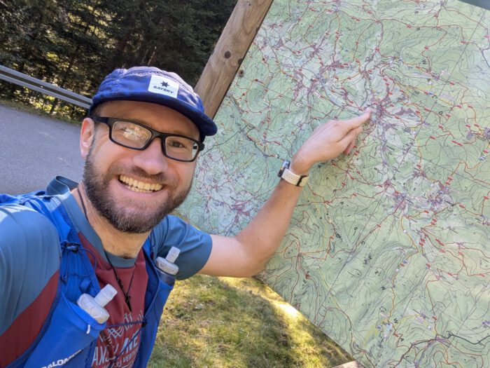 Lächelnder Thorsten mit Brille und Cap zeigt auf eine große Wanderkarte im Freien