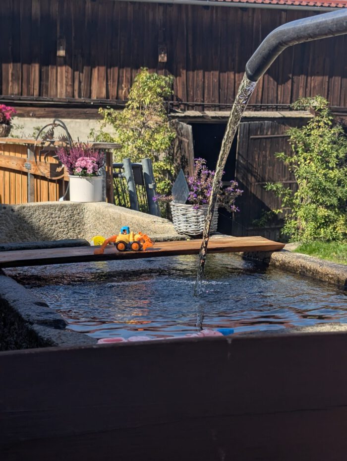 Spielzeugbagger auf einem Holzbrett über einem Steinbrunnen, in den Wasser aus einem gebogenen Rohr fließt