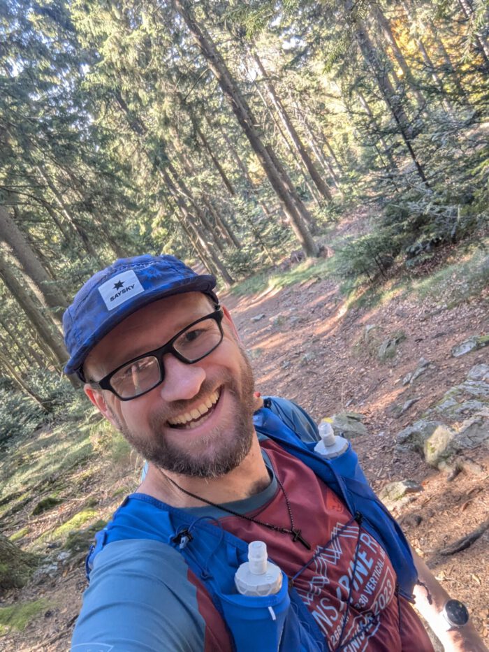 Lächelnder Thorsten mit Brille, Mütze und Laufweste macht ein Selfie auf einem sonnigen Waldweg