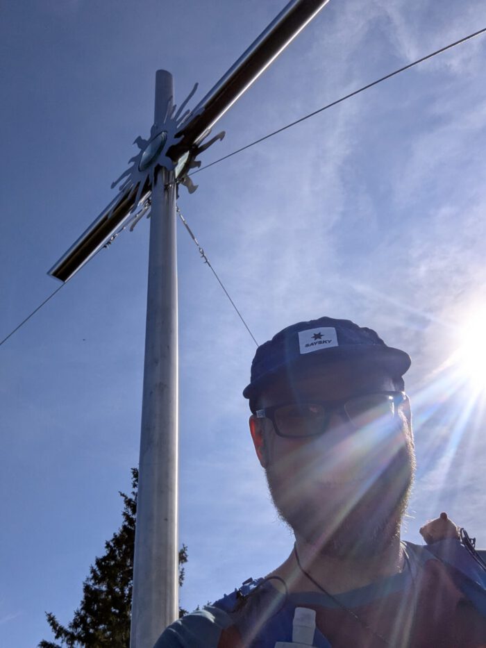 Thorsten mit Kappe und Brille macht ein Selfie vor einem metallenen Gipfelkreuz am Schwarzeck im Sonnenschein