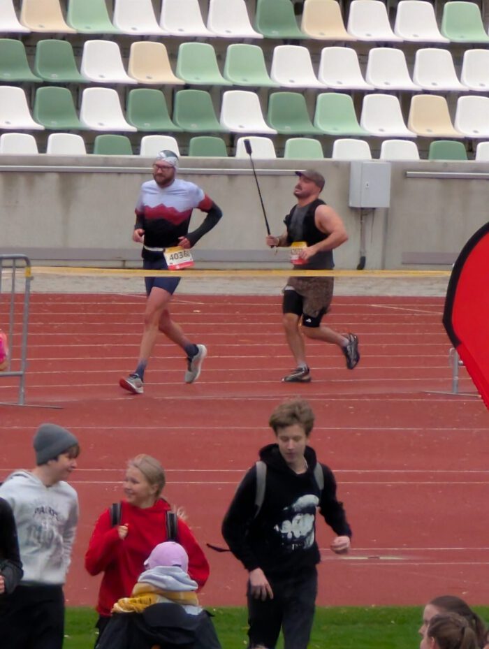 Zwei Läufer auf einer roten Laufbahn im Stadion vor leeren Tribünenrängen, im Vordergrund gehen Jugendliche